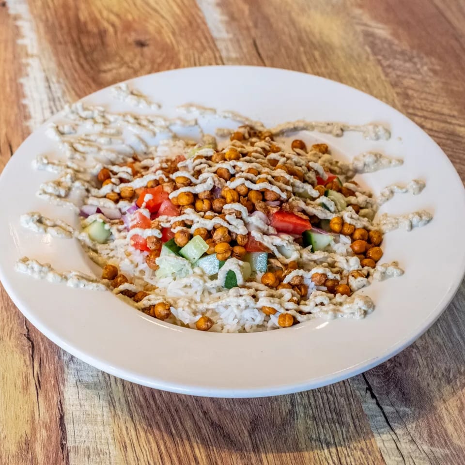 A white plate of rice topped with chickpeas, diced tomatoes, cucumbers, and a creamy sauce.