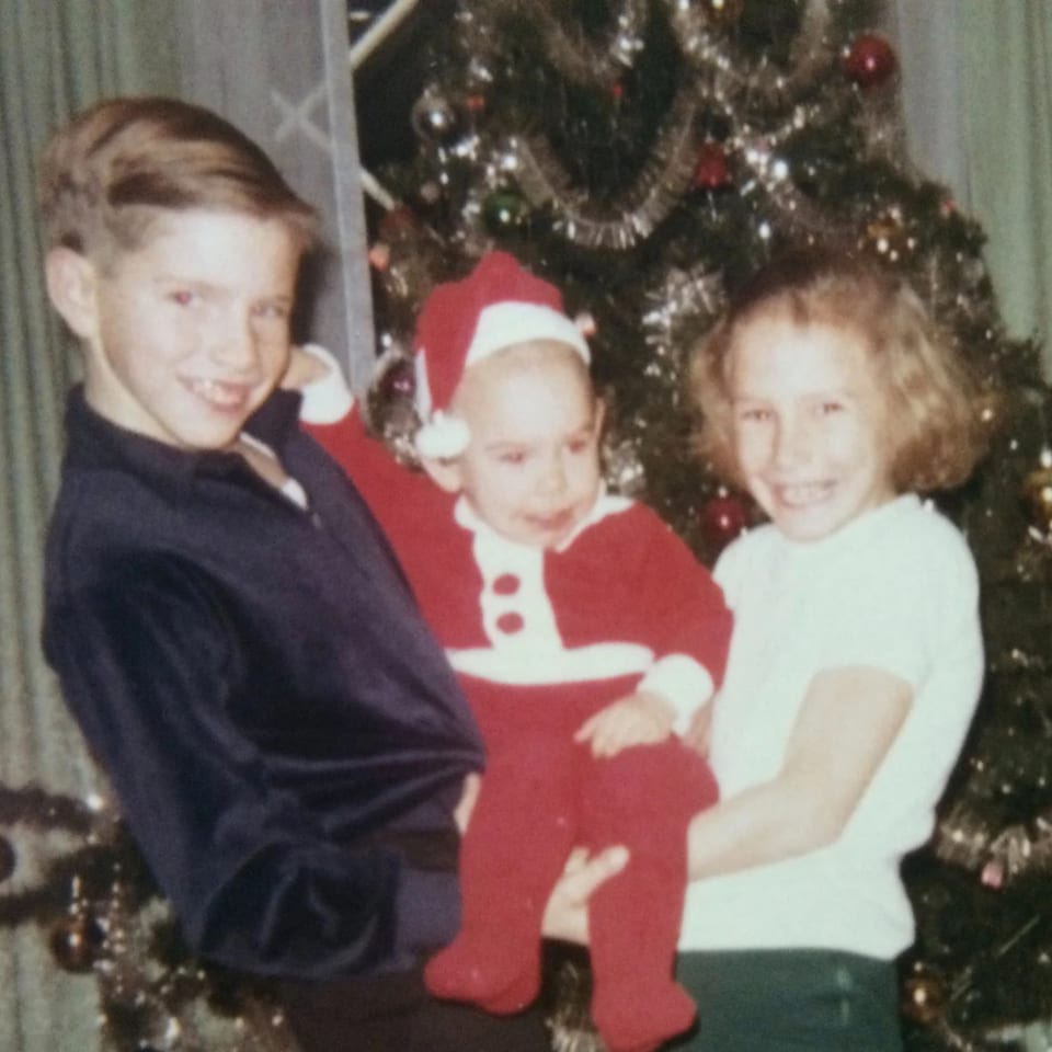 Three smiling children, one dressed as Santa, in front of a decorated Christmas tree.
